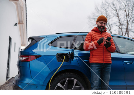 Man charging electric car during cold snowy day, using electric vehicle charging app, checking battery life, energy consumption on smart phone. Charging and driving electric vehicles during winter 110589591