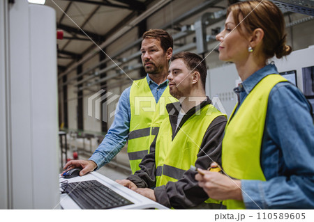 Young man with Down syndrome working in warehouse, colleague teaching him work with computer. Concept of workers with disabilities, support in workplace. 110589605