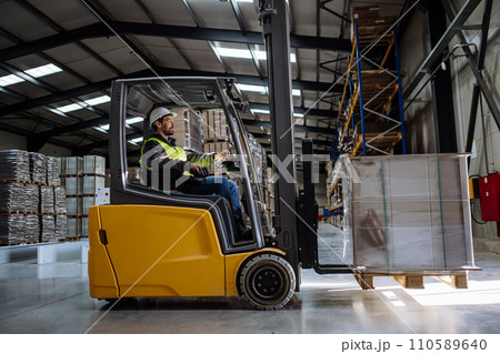 Side view of forklift in warehouse with male driver. Warehouse worker preparing products for shipmennt, delivery, checking stock in warehouse. 110589640