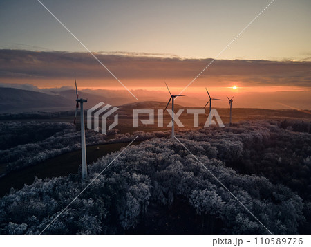 Aerial landscape photography of sunrise over frost-covered nature with wind turbines. Windmill in soft morning light with icy trees around. Concept of wind power as clean, renewable energy source. Aerial landscape photography of sunrise over frost-covered nature with wind turbines. Windmill in soft morning light with icy trees around. Concept of wind power as clean, renewable energy source. 110589726