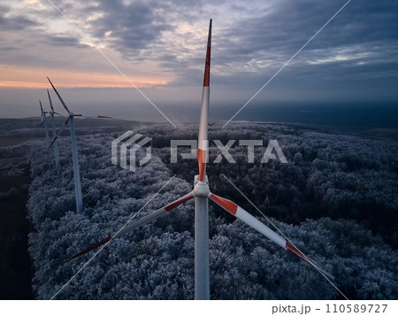 Aerial landscape photography of sunrise over frost-covered nature with wind turbines. Windmill in soft morning light with icy trees around. Concept of wind power as clean, renewable energy source. 110589727