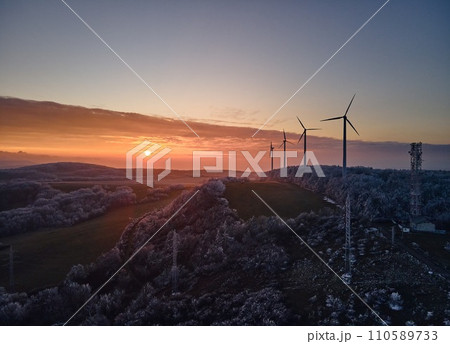 Aerial landscape photography of sunrise over frost-covered nature with wind turbines. Windmill in soft morning light with icy trees around. Concept of wind power as clean, renewable energy source. 110589733