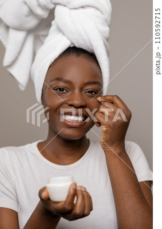 Joyful woman applying daily care cream after taking shower standing with towel on head 110592715