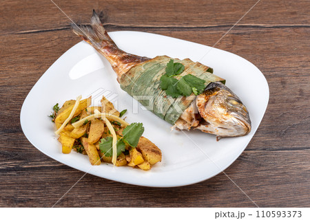Baked dorado fish in bamboo leaves, baked potatoes with bamboo shoots on a white plate. Baked dorado fish in bamboo leaves, baked potatoes with bamboo shoots on a white plate. 110593373