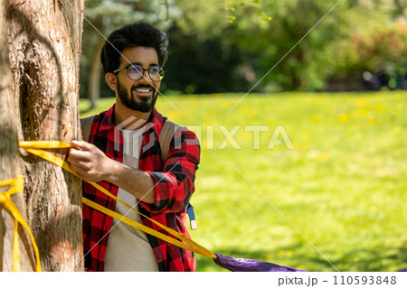 Man in checkered shirt tying ropes to the tree and adjusting hammock 110593848
