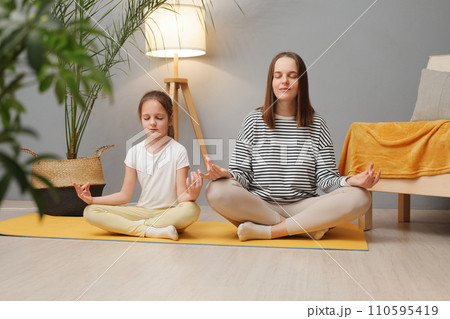 Family yoga. Beautiful young woman and her charming little daughter doing yoga together at home sitting in lotus position on floor in living room mom teaching child to meditate Family yoga. Beautiful young woman and her charming little daughter doing yoga together at home sitting in lotus position on floor in living room mom teaching child to meditate 110595419