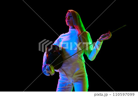 Portrait of young woman, fencer, athlete in white uniform posing with sword over black background in neon light 110597099