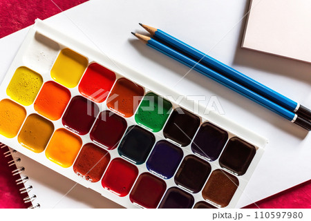 Colorful watercolor or acrylic paints and pencils on a white sheet of paper on red background. Design studio table top view. Colorful watercolor or acrylic paints and pencils on a white sheet of paper on red background. Design studio table top view. 110597980