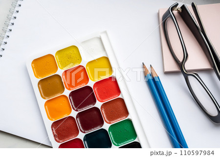 Colorful watercolor or acrylic paints, pencils and glasses on a white sheet of paper on a white background. Design studio table top view.  110597984