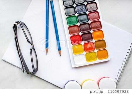 Colorful watercolor or acrylic paints, pencils and glasses on a white sheet of paper on a white background. Design studio table top view. Colorful watercolor or acrylic paints, pencils and glasses on a white sheet of paper on a white background. Design studio table top view. 110597988