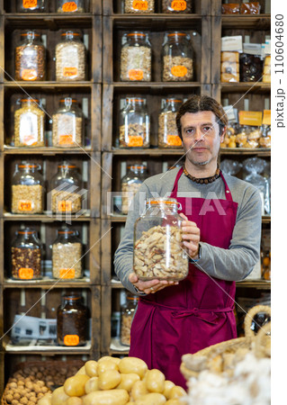 Male seller holding jar of brazil nuts standing in nut section of supermarket 110604680
