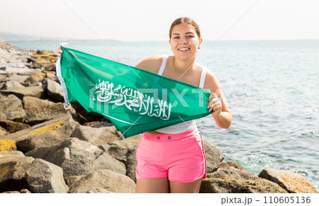 Portrait of positive young woman with the flag of Saudi Arabia on the beach on sunny day 110605136