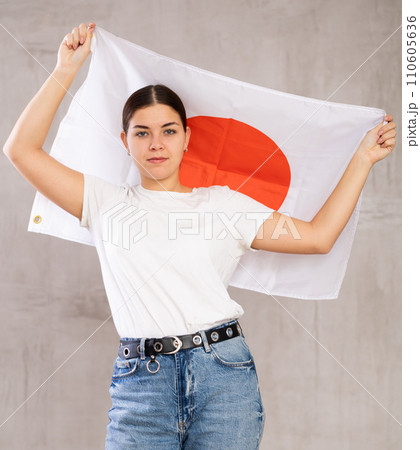 Serious young woman holding state flag of Japan against gray wall background indoors Serious young woman holding state flag of Japan against gray wall background indoors 110605636