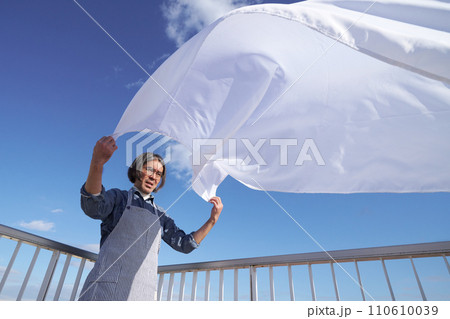 青空のもとで洗濯物のシーツを広げるミドル主夫 110610039