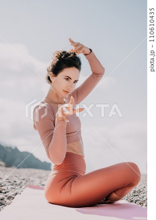 Middle aged well looking woman with black hair, fitness instructor in leggings and tops doing stretching and pilates on yoga mat near the sea. Female fitness yoga routine concept. Healthy lifestyle 110610435