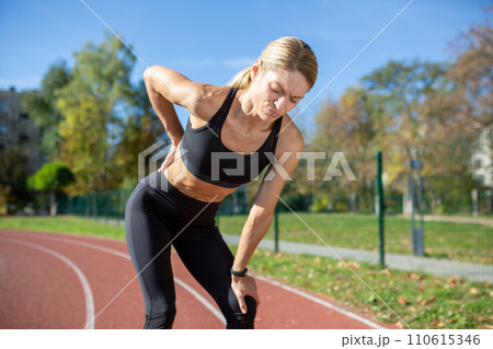 A fit woman in sports attire pauses with hands on knees, exhausted after a strenuous run on a sunny track. 110615346