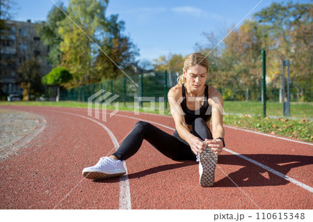 A determined woman stretches her leg muscles before a run on a track field, illustrating fitness and preparation. 110615348