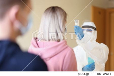 Person wearing protective medical suit and mask measures temperature with thermometer to visitors 110618984