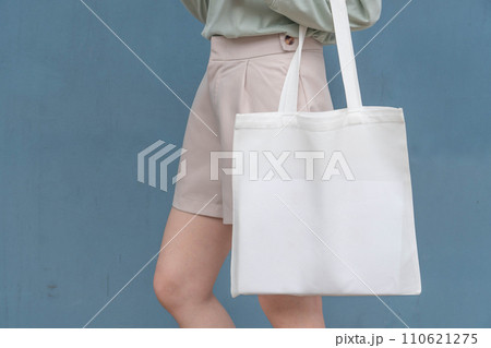 Blank white tote bag canvas fabric with handle mock up design. Close up of woman holding eco or reusable shopping bag against blue metal wall. No plastic bag and ecology concept. Blank white tote bag canvas fabric with handle mock up design. Close up of woman holding eco or reusable shopping bag against blue metal wall. No plastic bag and ecology concept. 110621275