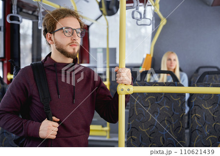 Close up of pensive adult male standing looking at window in public transport. Crop of cute young man in burgundy hoodie travelling with blur blonde sitting in urban transport. Concept of city life. Close up of pensive adult male standing looking at window in public transport. Crop of cute young man in burgundy hoodie travelling with blur blonde sitting in urban transport. Concept of city life. 110621439