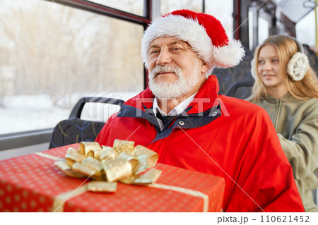 Front view of mature bearded male in red jacket smiling at camera and young girl sitting behind in earmuffs in public transport. Crop of passengers travelling by bus on holydays. Concept of joy. Front view of mature bearded male in red jacket smiling at camera and young girl sitting behind in earmuffs in public transport. Crop of passengers travelling by bus on holydays. Concept of joy. 110621452