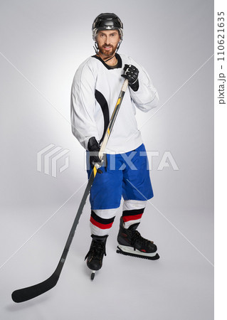 Smiling ice hockey player with stick and protective helmet posing in studio. Front view of handsome bearded hockey athlete looking at camera, isolated on gray background. Concept of winter sports. 110621635