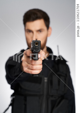 Armed caucasian police officer aiming with handgun to camera. Selective focus, close up of police pistol's muzzle, with blurred silhouette of policeman on background. Concept of weapon, target. Armed caucasian police officer aiming with handgun to camera. Selective focus, close up of police pistol's muzzle, with blurred silhouette of policeman on background. Concept of weapon, target. 110621704