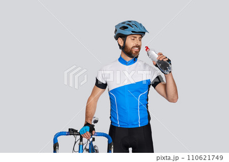 Muscular male cyclist in helmet drinking water, while resting in studio. Portrait of bearded athlete quenching thirst, while standing with bike, isolated on grey. Concept of sport activity, cycling. Muscular male cyclist in helmet drinking water, while resting in studio. Portrait of bearded athlete quenching thirst, while standing with bike, isolated on grey. Concept of sport activity, cycling. 110621749