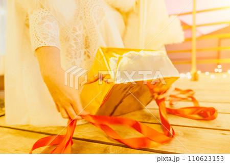 A woman in a white dress is holding a gold box with a red ribbon. She is wearing a crown on her head. The woman appears to be opening the gift box. 110623153