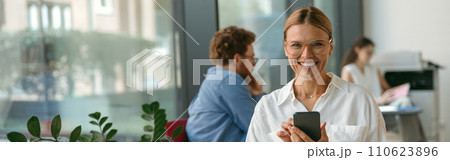 Woman freelancer use mobile phone while working on laptop in modern coworking and looks camera 110623896