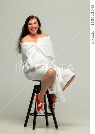 A beautiful middle-aged woman wrapped in a white sheet poses on a studio chair. A beautiful middle-aged woman wrapped in a white sheet poses on a studio chair. 110623948