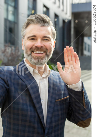 Smiling mature businessman in a stylish suit waving hello, standing outdoors by an office building, exuding leadership. Smiling mature businessman in a stylish suit waving hello, standing outdoors by an office building, exuding leadership. 110625674