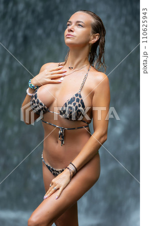 Beautiful young woman relaxes under a waterfall. Sexy girl in a bikini posing near a waterfall in the tropics. A traveler bathes under streams of water flowing from the mountains. Beautiful young woman relaxes under a waterfall. Sexy girl in a bikini posing near a waterfall in the tropics. A traveler bathes under streams of water flowing from the mountains. 110625943