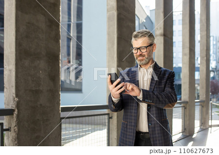 Smiling senior businessman in a stylish suit engaged with his smartphone outside a contemporary office setting. 110627673