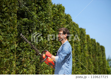 Smiling handyman in glasses trimming, shaping conifer hedge with modern electric trimmer at sunny day. Side view of happy, focused man pruning evergreen bushes outdoors. Concept of gardening services. Smiling handyman in glasses trimming, shaping conifer hedge with modern electric trimmer at sunny day. Side view of happy, focused man pruning evergreen bushes outdoors. Concept of gardening services. 110627725