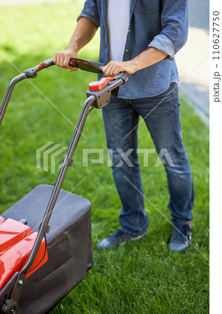Unrecognizable male gardener pushing modern lawn mower, while walking on grass. Above view of handyman in denim taking care of turf with grass trimmer outdoors. Concept of garden equipment. 110627750