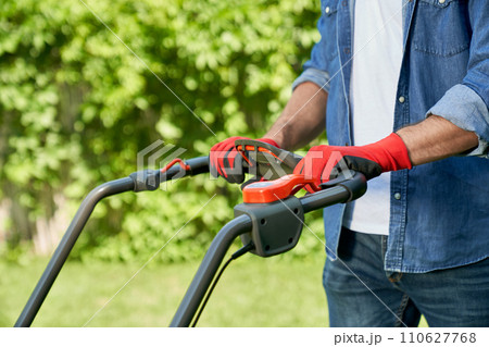 Crop view of gardener in casual outfit mowing lawn with modern lawn mower in summer. Close up view of muscular male hands in gloves on lawn mower handle against hedge background. Concept of gardening. 110627768