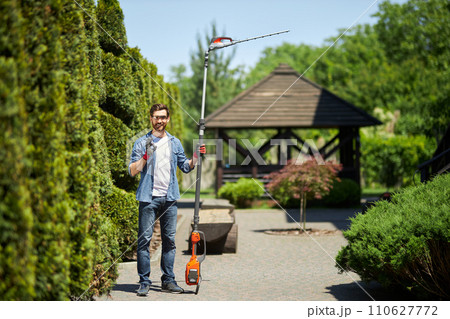 Happy male worker showing okay sign, while standing with electric hedge trimmer outdoors. Front view of smiling gardener with bush cutter gesturing, while looking at camera. Concept of work, hobby. Happy male worker showing okay sign, while standing with electric hedge trimmer outdoors. Front view of smiling gardener with bush cutter gesturing, while looking at camera. Concept of work, hobby. 110627772