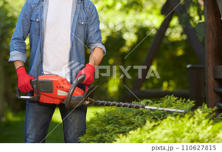Unrecognizable strong male gardener trimming conifer hedges with modern electric loppers outdoors. Crop view of man in gloves pruning bushes with pro equipment in garden. Concept of gardening. 110627815