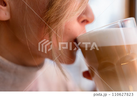 Woman with blonde hair sips cappuccino in a cafe. She is holding the glass up to her face, taking a sip of the drink. Woman with blonde hair sips cappuccino in a cafe. She is holding the glass up to her face, taking a sip of the drink. 110627823