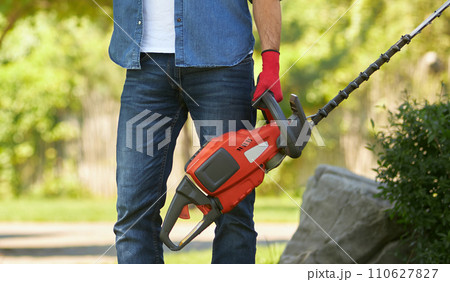 Unrecognizable muscular man in jeans standing with modern electric hedge trimmer outdoors. Crop view of strong male worker holding equipment for pruning, on blurred background. Concept of gardening. Unrecognizable muscular man in jeans standing with modern electric hedge trimmer outdoors. Crop view of strong male worker holding equipment for pruning, on blurred background. Concept of gardening. 110627827