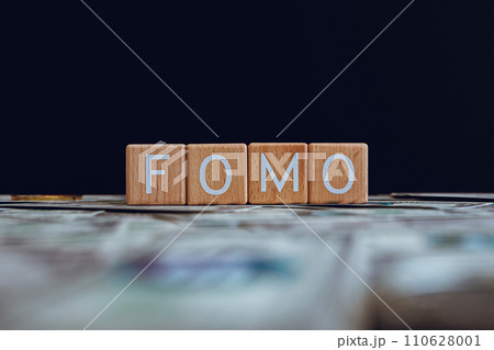 Wooden blocks with the text "FOMO" on a black background and crypto banknotes scattered on the ground. Wooden blocks with the text "FOMO" on a black background and crypto banknotes scattered on the ground. 110628001