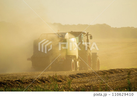 Combine harvester pressing straw from field into bales driving field on sunny summer evening. Field with bales of pressed wheat. Lots of dust on field. Agricultural agro-industrial harvesting works 110629410