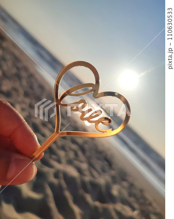 Person holding in fingers hand stick in shape golden heart and word Love on background sea and sea waves in seashore on sunny summer day close-up. Concept love romance amour St Valentines Day Person holding in fingers hand stick in shape golden heart and word Love on background sea and sea waves in seashore on sunny summer day close-up. Concept love romance amour St Valentines Day 110630533