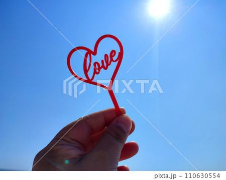 Person holding stick in shape red heart and word Love in hand on background of blue sky with shining sun on sunny summer day. Concept love relationship romance amour celebration St Valentines Day 110630554