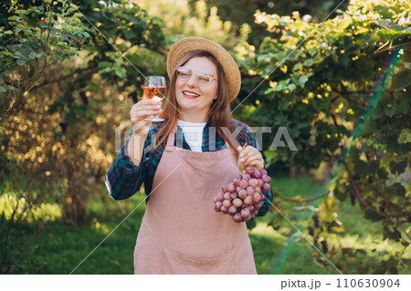 Beautiful young woman in hat with glass of pink wine in vineyard on sunny day. Food and drink concept, small local farming. 30s woman shows a pink grapes harvested Beautiful young woman in hat with glass of pink wine in vineyard on sunny day. Food and drink concept, small local farming. 30s woman shows a pink grapes harvested 110630904