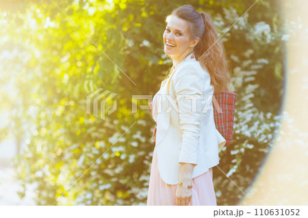 smiling trendy woman in pink dress and white jacket in city 110631052