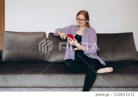 Young smiling woman holding smartphone, using cellphone modern technology, checking cell phone apps, texting, watching video, browsing internet for shopping. Girl sitting on sofa at home Young smiling woman holding smartphone, using cellphone modern technology, checking cell phone apps, texting, watching video, browsing internet for shopping. Girl sitting on sofa at home 110631211