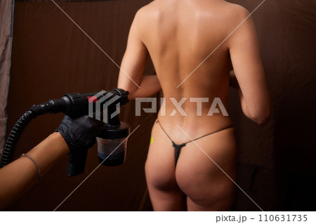 Cosmetologist using airbrush for spray tan apply to young woman body in beauty salon. 110631735