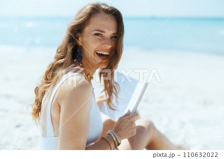 smiling stylish woman on beach with straw bag and book 110632022